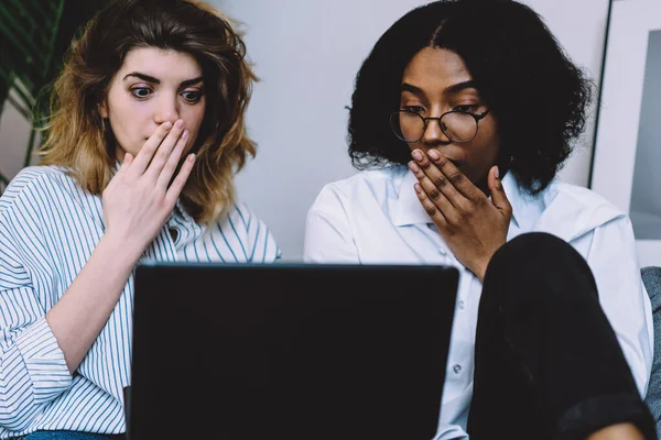 Astonished caucasian and african american young women shocked with last publication on web page on common website sitting at laptop device at home interior.Amazed multicultural friends reading news