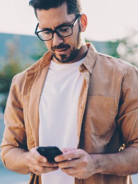 Görme düzeltmesi için yetişkin bir adam cep telefonunda uygulamayı güncellerken şehir ortamında ayakta duruyor ve internet bağlantısı kullanıyor, Kafkas erkek blogcu mesaj yazıyor