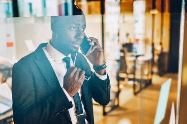 Deneyimli Afro-Amerikan girişimci resmi giyinmiş akıllı telefon cihazıyla konuşuyor cam ofis duvarının arkasında duruyor. Koyu tenli gururlu CEO telefonla arıyor.