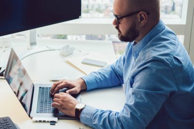 Modern ofiste bilgisayarlı ve tabletli dizüstü bilgisayar kullanan odaklanmış kel adamın yan görüntüsü.
