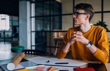 Gündelik giyinmiş hippi adam masaüstünde ekipman ve çizimlerle otururken yüzünü çeviriyor. Stüdyoda çalışarak tasarım projesi üzerine düşünen düşünceli genç adam.