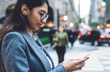 Odaklanmış, yuvarlak gözlüklü, gri montlu, New York 'un kalabalık caddesinde, bulanık arka planda yaya geçidinde akıllı telefondan konuşan esmer bir kadının yan görüntüsü.