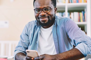 Neşeli Afrikalı Amerikalı 'nın elinde cep telefonuyla dizüstü bilgisayarla oturarak günlük giysiler içinde gözlük taktığını düşünmek.