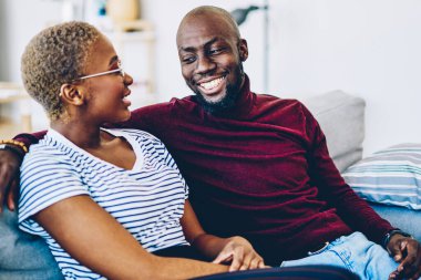 Pozitif Afro-Amerikan hipster çifti birbirlerine bakıyor ve birlikte vakit geçiriyorlar, gülümseyen esmer tenli adam karısına sarılıyor oturma odasında boş zamanlarında sohbet ediyorlar.