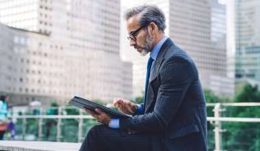 Gözlüklü ve siyah kostümlü yakışıklı iş adamının New York 'ta bulanık şehir geçmişinde tablet kullanmasının yan görüntüsü...