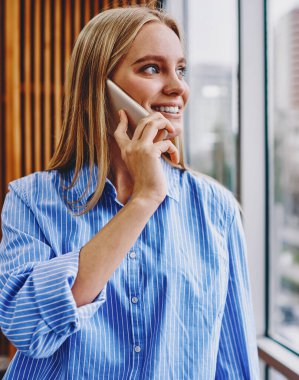 Akıllı telefon cihazından arkadaşınla iletişim kurarken camdan dışarı bakan mutlu, genç bir kadın. Ofisin içindeki modern selülomu çağırırken gülen olumlu hipster kızı.