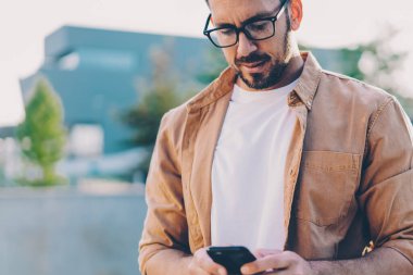 Görme düzeltmesi için yetişkin bir adam cep telefonunda uygulamayı güncellerken şehir ortamında ayakta duruyor ve internet bağlantısı kullanıyor, Kafkas erkek blogcu mesaj yazıyor