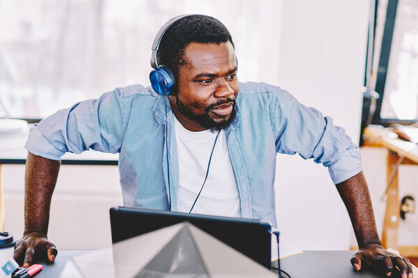 Serious African American man in casual clothes looking away and wearing headphones while sitting at table with laptop in modern office