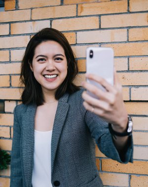 Sıradan giysiler içinde gülümseyen genç bir kadın binanın tuğla duvarına yaslanmış kameraya bakarken akıllı telefonuyla selfie çekiyor.