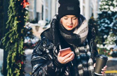 Kışlık kıyafetleri içinde, elinde siyah bardakla akıllı telefon kullanan ve karlı caddede Noel ağaçlarının yanında duran konsantre genç bayan.