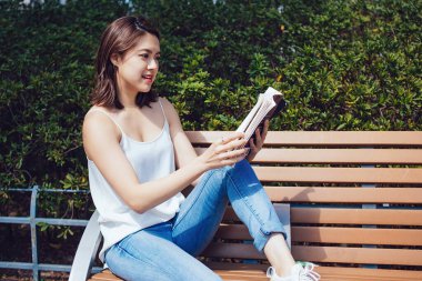 Kotlu rahat bir etnik kadın yazın güneşli bir parkta ahşap bankta oturup ilginç bir kitap okuyor.