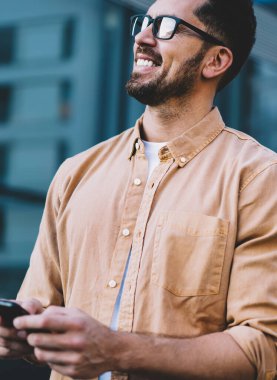 Dijital teknolojinin neşeli kullanıcıları ellerinde cep telefonuyla şehir ortamında dikiliyor ve gülümsüyor, sıradan giyinen mutlu beyaz adam akıllı telefon aleti tutuyor ve eğleniyor.