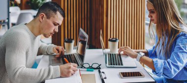 Gündelik giysiler giyen iki yetenekli öğrenci, modern ofiste kablosuz internet bağlantısı kullanarak dizüstü bilgisayarların başında sınavlara hazırlanıyor.