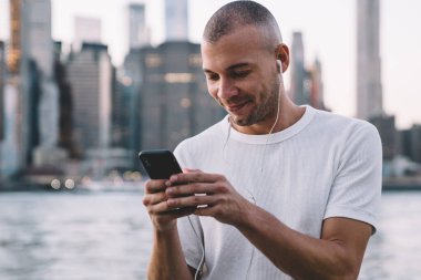 Genç gülümseyen etnik adam kulaklıkla müzik dinlerken akıllı telefon kullanıyor ve nehir ve şehrin bulanık arka planına karşı korkuluklara yaslanıyor.