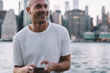 Beyaz tişörtlü, gülümseyen, kulaklıkla müzik dinleyen ve nehir kenarındaki metal tellere yaslanmış akıllı telefon tutan genç bir adam.