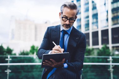 New York binalarının arka planında otururken Stylus 'la tablet kullanan, iş elbisesi ve gözlük takan yakışıklı, orta yaşlı adam.