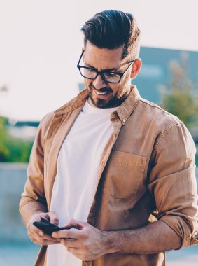 Optik gözlüklü başarılı adam şehir ortamında boş zamanlarında mobil uygulama tebligatını kontrol ediyor, gündelik tişört giymiş mutlu erkek çevrimiçi iletişim sırasında SMS mesajı gönderiyor