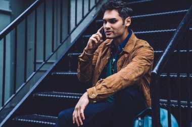 Yakışıklı, rahat giyinen, dijital akıllı telefonla konuşan, aşağı bakan ve koyu demir merdivenlerde oturan sakin bir erkek.