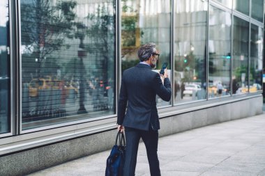 New York 'ta akıllı telefon ve çantayla şehir caddesinde yürüyen şık, koyu renk takım elbiseli başarılı orta yaşlı bir adamın arka planı.