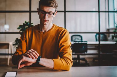 Gözlüklü genç adam modern saate bakıyor ve masasının içindeki kafeteryada oturan aleti kontrol ediyor. Günlük kıyafet giymiş konsantre hipster adam toplantı saatini bekliyor.
