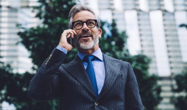 Modern dijital akıllı telefonla konuşan, konuşan, iletişim kuran, gülümseyen ve şehir geçmişine bakan resmi giysili, gri saçlı mutlu orta yaşlı erkek.