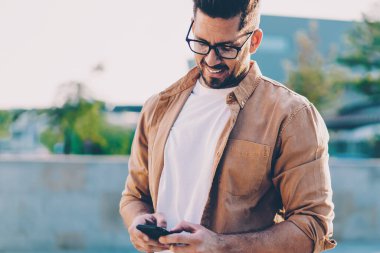 Optik gözlüklü başarılı adam şehir ortamında boş zamanlarında mobil uygulama tebligatını kontrol ediyor, gündelik tişört giymiş mutlu erkek çevrimiçi iletişim sırasında SMS mesajı gönderiyor