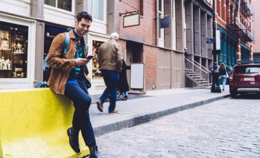 Neşeli stil sahibi genç bir erkek modern dijital aletlerle geziyor ve iletişim kuruyor. Oturuyor, gülümsüyor ve şehirdeki sokaklara bakıyor.