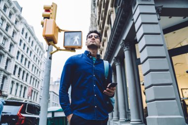 Gündüz vakti şehir kavşağında duran, rahat gömlekli, gözlüklü, akıllı telefon ve sırt çantalı, kendinden emin, etnik, yetişkin bir adam.