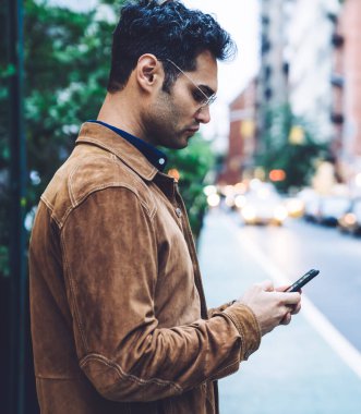 Ceketli ve gözlüklü, akıllı telefon kullanan, güpegündüz trafik şeridinin yanında duran kendinden emin, yakışıklı, esmer bir adam.