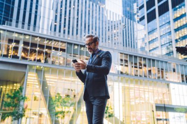 Koyu takım elbiseli, beyaz saçlı ve gözlüklü akıllı iş adamı New York 'ta şehir planında akıllı telefondan mesaj atıyor.