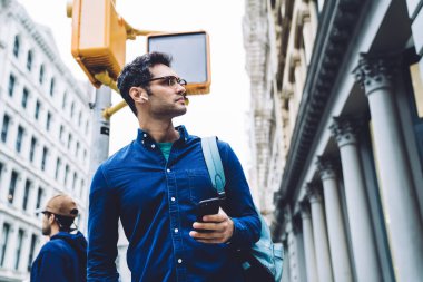 Kot gömlekli, kulaklıklı ve akıllı telefonlu gözlüklü yakışıklı yetişkin bir adamın gündüz vakti sokakta dikilirken etrafa bakınmasından.