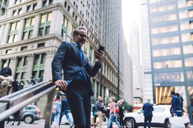 Aşağıdan gülümseyen orta yaşlı gözlüklü ve modaya uygun bir iş adamı ve New York 'ta işlek bir metropol sokağındaki tırabzanlara dayanan akıllı telefonlu.