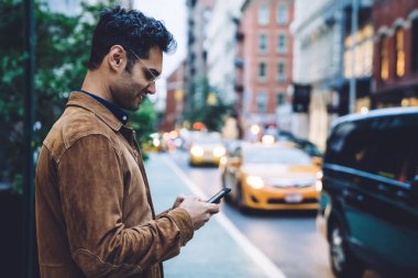 Ceketli ve gözlüklü yakışıklı, esmer adamın gündüz şeridinin yanında dururken akıllı telefon kullanışının yan görüntüsü.