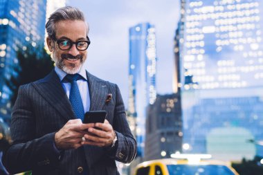Gözlüklü, takım elbiseli, telefonda yazan ve gülümseyen orta yaşlı yakışıklı bir erkek. New York karayolu ve gökdelenlerin arka planında dikilirken.