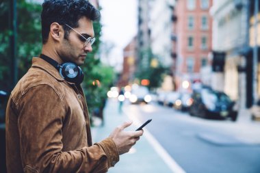 Somurtkan etnik esmer, ceketli ve kulaklıklı, akıllı telefon kullanan, güpegündüz trafik şeridinin yanında duran yakışıklı, esmer adamın yan görünüşü.