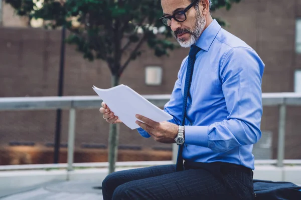 Side view of middle aged man in business suit and glasses sitting on bench and analyzing papers on New York street