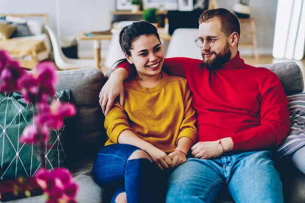 Positive couple in love spending time together at comfortable apartment talking to each other on couch, happy romantic woman and man enjoying leisure at home interior communicating and smiling