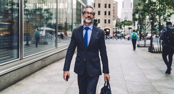 Successful content classy man in trendy suit and sunglasses holding smartphone and bag in hands while walking confidently on New York street