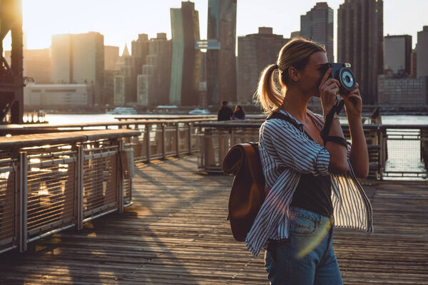 Young female tourist with rucksack taking pictures of evening city - New York using vintage instant camera for capture moment of summer American vacations, concept of amateur photographing