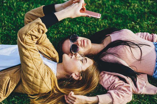 Young female friends lying on green grass together in trendy sunglasses using smartphone camera for selfie, pretty hipster girls in stylish wear taking picture on mobile phone resting in park