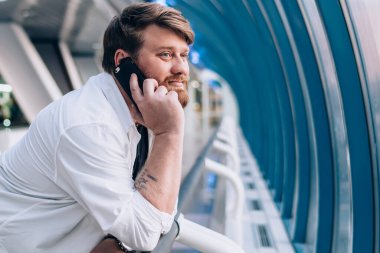 Hafif geometrik koridorda çitlere yaslanmış, cep telefonuyla konuşan hoş sakallı adamın yan görüntüsü.