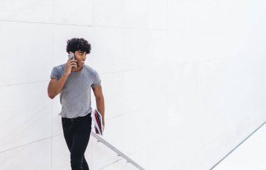 Beyaz iş merkezinde merdivenleri çıkarken akıllı telefondan konuşan neşeli, modern bir etnik adam. Elinde kağıtlar, başka tarafa bakıyor.