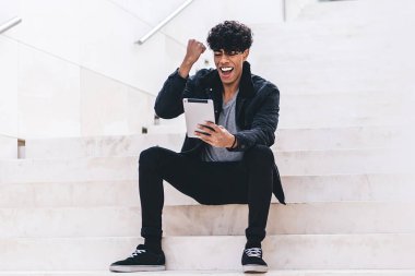 Gündüz vakti şehir merdivenlerinde otururken günlük kıyafetleriyle tablet üzerinde gezinen şık, genç, esmer, afro-amerikan bir adam.