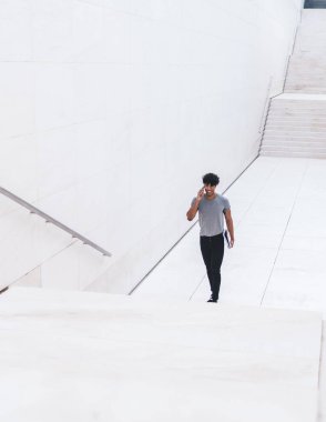 Zeki telefonuyla konuşan, elinde belgelerle beyaz alışveriş merkezinde yürürken gülümseyen, sıradan giyinen neşeli etnik adam.