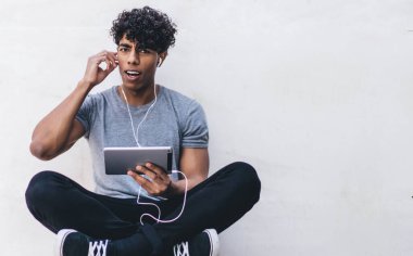 Şaşkın, genç, şaşkın, sıradan giyinen siyah erkek. Tablet ve kulaklıklarla bacak bacak bacak üstüne atarak dışarıdaki kameraya bakıyor.