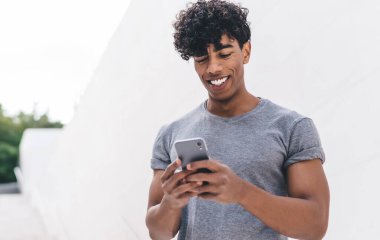 Gülümseyen, kıvırcık, gri tişörtlü genç adam beyaz duvarın önündeki merdivenlerde geziyor.