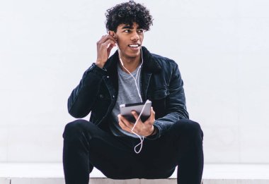 Tabletle merdivenlerde oturmuş kulaklıkla müzik dinlerken beyaz duvara bakan Afro-Amerikalı genç yakışıklı bir adam.