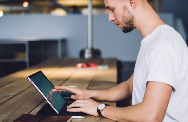 Odaklanmış profesyonel hippi yetişkin işçi ahşap masada oturmuş çatı katında dokunmatik ekranlı bir defter kullanıyor.