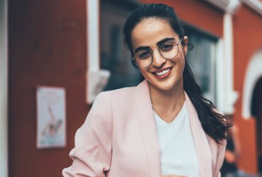 Gözlüklü, beyaz gömlekli, pembe ceketli, kendine güveni tam bir siyah kadın sokakta dikilirken kameraya gülümseyen ve bulanık renkli dış görünüşlü kadınlara karşı akıllı telefon kullanıyor.