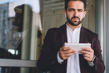 Takım elbiseli asık suratlı etnik adam modern binanın penceresinin yanındaki tableti incelerken kameraya bakıyor ve müzik dinliyor.